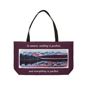 May include: A maroon tote bag with black handles. The bag features a landscape painting of mountains, a lake, and trees. Text on the bag reads, "In nature, nothing is perfect, and everything is perfect."