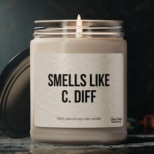 May include: A white soy wax candle in a glass jar with a black lid. The label reads "Smells Like C. Diff" and "100% natural soy wax candle".