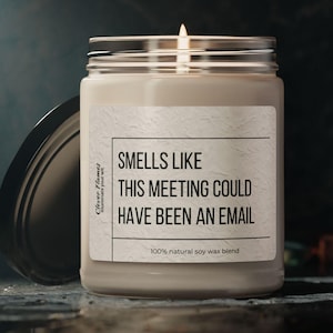 Peut inclure: Une bougie en pot de verre transparent avec une mèche allumée et un couvercle noir posé à côté. L'étiquette blanche porte le texte : Clever Flames, SMELLS LIKE THIS MEETING COULD HAVE BEEN AN EMAIL, et 100% natural soy wax blend en noir.