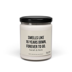 May include: A white candle jar with a black lid. The label reads "Smells like 16 years down, forever to go. Sarah & Nick 100% natural soy wax blend".