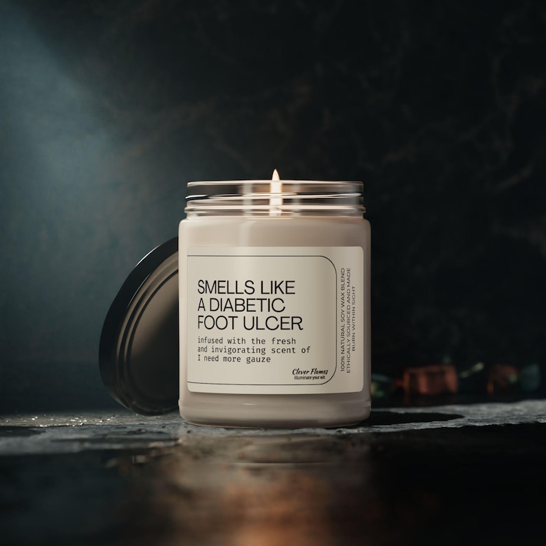 A lit white candle in a glass container labeled Smells Like a Diabetic Foot Ulcer sits on a dark surface, surrounded by unique jewelry. The dimly lit background adds to the moody atmosphere, as if adorning the scene with mysterious elegance.