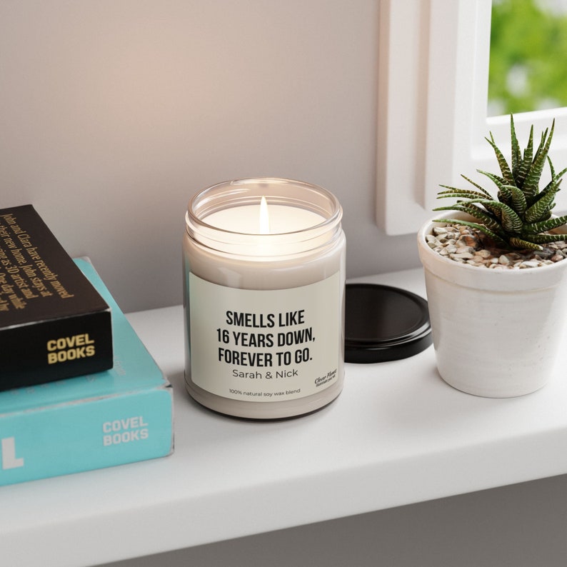 May include: A white candle with a black label that reads "Smells like 16 years down. Forever to go. Sarah & Nick" and "100% natural soy wax blend". The candle is lit and sitting on a white surface.
