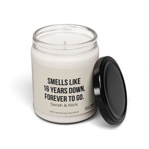 May include: A white candle in a glass jar with a black lid. The label reads "Smells like 16 years down, forever to go. Sarah & Nick 100% natural soy wax blend".