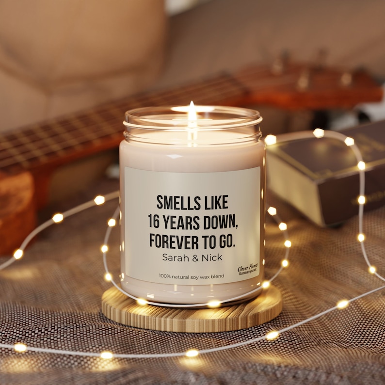 May include: A white candle with a black label that reads "Smells like 16 years down, forever to go. Sarah & Nick 100% natural soy wax blend". The candle is sitting on a wooden coaster.