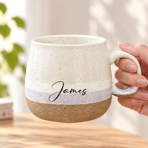 May include: A ceramic mug with a speckled cream top, a light gray middle band, and a textured brown base. The name James is printed in black cursive script on the middle band. The mug features a rounded body and a curved handle on the right side.