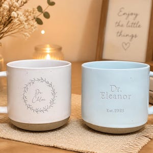 May include: Two ceramic mugs with speckled finishes and handles. The white mug features a gray botanical wreath surrounding the text Dr. Chloe. The light blue mug is embossed with the text Dr. Eleanor and Est. 2025. Both mugs have a solid gray base.