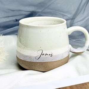 Puede incluir: Taza de cerámica color crema con forma redondeada y asa grande. La taza presenta un acabado moteado y un diseño de bloques de color con una banda gris claro y marrón. El nombre "James" está escrito en cursiva en la taza.