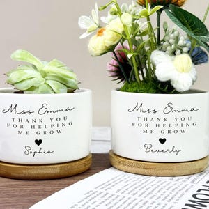 May include: Two white ceramic plant pots with bamboo saucers. One holds a succulent, the other a floral arrangement. Each pot has the text "Miss Emma, THANK YOU FOR HELPING ME GROW" with a name below. The succulent pot is labeled "Sophia", the floral pot is labeled "Beverly".