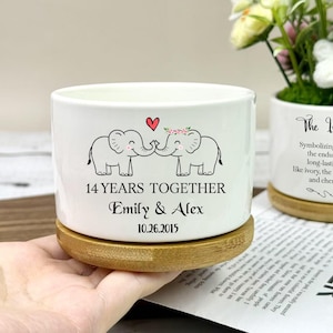May include: A white ceramic planter with a bamboo base. The planter features a design of two elephants touching trunks, a heart, and the text "14 YEARS TOGETHER Emily & Alex 10.26.2015". A second planter with text is also visible.