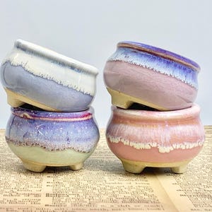 Puede incluir: Cuatro pequeñas macetas de cerámica apiladas en tonos azules, morados y rosas. Cada maceta tiene una forma redondeada con un acabado texturizado y esmaltado y se asienta sobre tres pequeños pies. Adecuado para plantas pequeñas o suculentas.