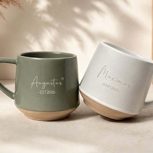 Può includere: Due tazze in ceramica con manico e base in argilla naturale. La tazza verde salvia riporta la scritta Augustus e EST 2026. La tazza bianco sporco riporta la scritta Maximus e 04.05.2026. Entrambe le tazze presentano un piccolo cuore vicino al nome.