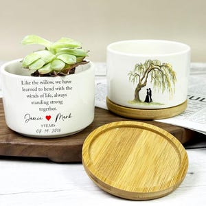 May include: Two white ceramic planters with a succulent plant and a wooden base. One planter has a willow tree illustration and the other has a quote and names. A wooden saucer is in the foreground.