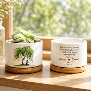 May include: Two white ceramic cylindrical planters with bamboo saucers. One planter features a watercolour illustration of a willow tree with two figures beneath it. The other planter displays the text: Like the willow, we have learned to bend with the winds of life, always standing strong together. Vanessa, a red heart, Daniel, and the date 13.08.2017.