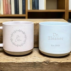 May include: Two ceramic mugs with handles. One is light pink with "Dr. Chloe" inside a wreath. The other is light blue with "Dr. Eleanor Est. 2025". Both mugs have a speckled finish and a tan base, set on a wooden surface.