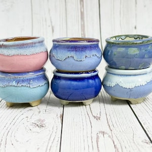 Può includere: Sei piccoli vasi in ceramica nei toni del blu, rosa e bianco sono impilati su una superficie di legno chiaro. Ogni vaso ha una forma arrotondata con una finitura strutturata e smaltata. I vasi sono adatti per piccole piante.