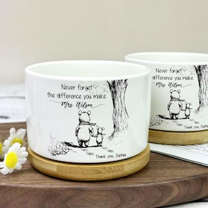 Può includere: Due fioriere in ceramica bianca con base in bambù. Ogni fioriera presenta un'illustrazione in bianco e nero di un orso e di un animale più piccolo, con il testo "Never forget the difference you make Mrs. Wilson" e "Thank you, Sophia."