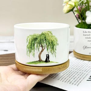 May include: White ceramic planter featuring a watercolour illustration of a weeping willow tree and a couple. The planter sits on a round bamboo base. The design includes the text "Like the willow, we have learned to bend with the winds of life, always standing strong together."