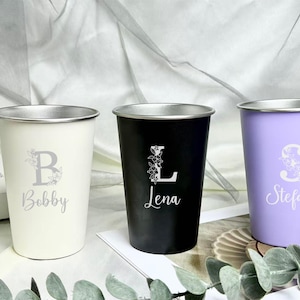 Puede incluir: Varios vasos personalizados de acero inoxidable en colores como amarillo, blanco roto, negro y lavanda. Cada vaso presenta un diseño floral con una letra y un nombre, perfecto para regalar.
