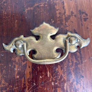 May include: A vintage brass drawer pull with an ornate design. The pull features a central handle and decorative elements on either side. The brass has a warm, aged patina, suggesting its antique nature. The background is a dark, wooden surface.