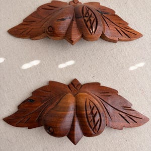 May include: Two intricately carved wooden decorative elements. Each element features a central fruit-like shape with two side leaves. The wood has a rich, dark brown color, and the leaves have detailed veining.