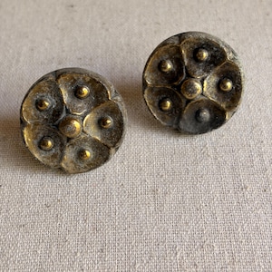 May include: Two antique brass-toned cabinet knobs. Each knob is round with a floral design featuring raised petal-like shapes and small, central, gold-colored accents. The knobs have a weathered, vintage appearance.