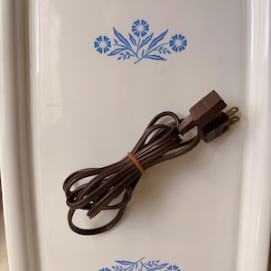 May include: A white rectangular tray with blue floral designs at the top and bottom. A brown electrical cord with a two-prong plug is coiled on the tray, secured with a rubber band. The tray has a glossy finish.