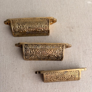 May include: Three antique brass drawer pulls with ornate floral and geometric designs. Each pull has a rectangular body with curved edges and small mounting tabs. The pulls are arranged vertically against a neutral background.