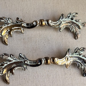 May include: Two matching metal drawer pulls featuring an ornate, elongated design with scrolling leaf-like motifs. The hardware has a distressed finish, combining metallic gold tones with areas of white paint in the recessed sections.