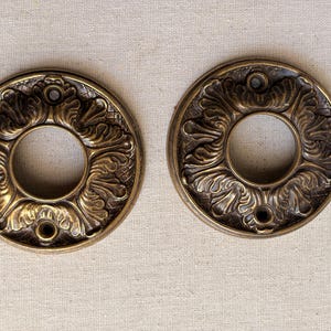 May include: Two antique brass door rosettes with ornate floral designs. Each rosette features a central circular opening and three screw holes. The rosettes have a warm, aged patina, suggesting they are vintage or antique.