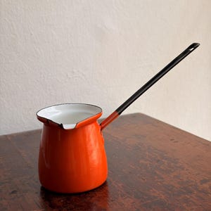 May include: An orange enamel coffee pot with a long black handle. The pot has a white interior and a black rim. The handle is attached to the side of the pot and extends outwards. The pot is sitting on a wooden surface.