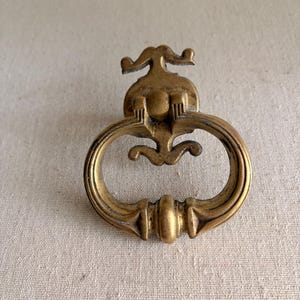 May include: Ornate brass drawer pull with a decorative design. The pull features a curved handle and an elaborate backplate with scroll-like details. The brass has a warm, aged patina, suggesting vintage or antique origins. Ideal for furniture restoration or adding a touch of elegance.
