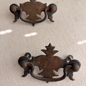 May include: Two antique bronze drawer pulls with ornate floral designs. Each pull features a central decorative element with a star-like top, a curved handle, and two mounting screws. The pulls have a weathered, aged appearance.