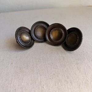 May include: Four circular cabinet knobs with a dark bronze finish. Each knob features a recessed center with a lighter, burnished gold tone and a decorative, textured border along the outer edge, creating a consistent look across all four pieces.