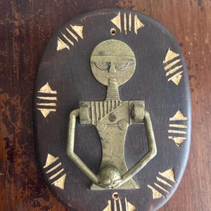 May include: An oval-shaped, dark brown wooden wall hanging with a brass figure in the center. The figure has a circular head, a long neck, and outstretched arms. Gold-colored arrow-like shapes surround the figure.