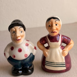 May include: A pair of vintage ceramic salt and pepper shakers. One shaker is a man with a black hat, white shirt with red polka dots, and blue pants. The other is a woman in a red dress and white apron.