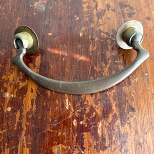 May include: A vintage-style drawer pull with a curved handle. The pull has a dark bronze finish and is attached to the cabinet with two circular brass-colored mounts. The hardware is designed for furniture restoration or adding a classic touch to cabinetry.