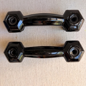 May include: Two black, glossy cabinet handles. Each handle has a curved center section and hexagonal ends with a central hole for mounting. The handles are likely made of a durable material and are designed for easy installation on furniture.