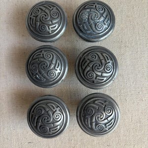 May include: Six antique-style, silver-toned cabinet knobs. Each knob features a raised, swirling Celtic knot design in the center. The knobs are round and have a slightly domed shape, suggesting they are made of metal.