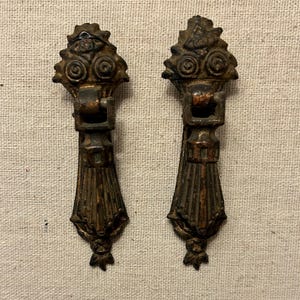 May include: Two antique bronze drawer pulls. Each pull features a decorative top with scrollwork and a rectangular handle. The lower portion has a fluted design, and a floral accent at the bottom. The pulls are aged with a patina.