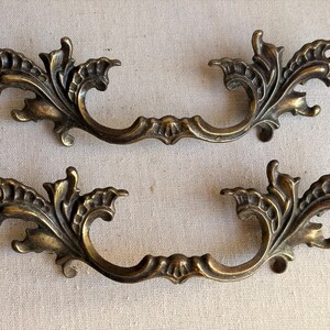 May include: Two antique bronze-toned drawer pulls with ornate, symmetrical floral and scroll designs. Each pull features a curved handle and decorative end pieces, perfect for adding a vintage touch to furniture. The pulls are displayed on a neutral background.
