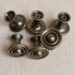 May include: A set of six round metal cabinet knobs with an antique bronze finish. The knobs feature a tiered, circular design with a central dome and a flared base. Three knobs are positioned to show their flat tops, while three show their threaded back.