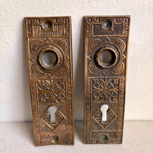 May include: Two rectangular bronze-toned metal door plates featuring intricate embossed geometric and floral patterns. Each plate includes a circular central opening for a knob, a lower keyhole cutout, and two screw holes at the top and bottom.