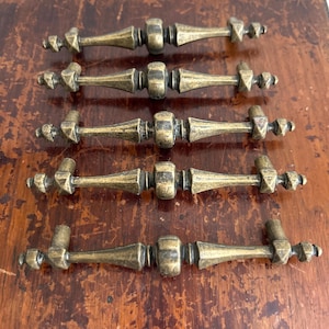 May include: Five antique brass drawer pulls with a detailed, ornate design. Each pull features a central barrel shape with tapered ends and decorative accents. The pulls are arranged on a wooden surface.