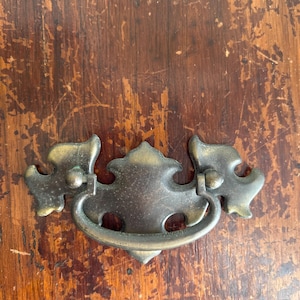 May include: Antique brass drawer pull with a decorative design. The pull features a curved handle and ornate details, perfect for vintage furniture restoration. The hardware is mounted on a distressed wooden surface.