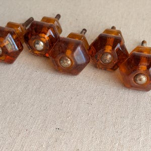 May include: A collection of amber-colored, faceted glass cabinet knobs with silver-toned metal accents. The knobs have a hexagonal shape and are arranged on a neutral-toned fabric surface. These vintage-style knobs are suitable for furniture restoration.