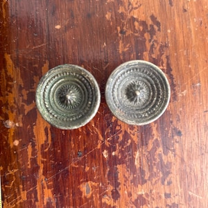 May include: Two antique, circular metal drawer pulls with a detailed, concentric design. The pulls have a tarnished, aged appearance, suggesting they are vintage or vintage-inspired. They are set against a warm, wooden surface.