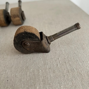 May include: Close-up of antique furniture casters. The casters have a cylindrical wheel made of a light brown material, likely wood or cork, with a dark rim. The metal frame and stem are a dark, aged color.