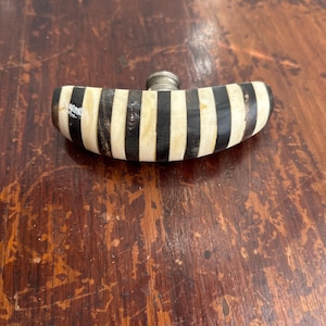 May include: A small, decorative flask with a curved, striped body. The flask features alternating black and cream-colored stripes. A silver-colored metal cap is attached to the top. The flask is resting on a wooden surface.