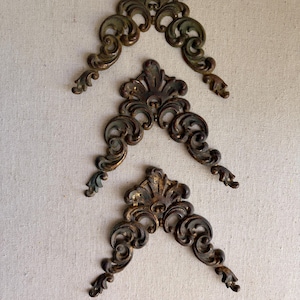 May include: Three matching decorative metal corner accents featuring an intricate scrollwork design with a dark, weathered bronze finish. Each piece displays a symmetrical pattern with curved flourishes and a central leaf-like motif at the top.