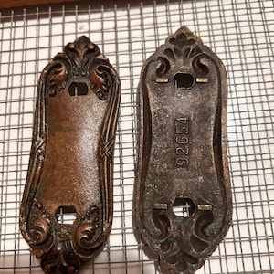 May include: Two antique bronze door plates with ornate floral designs. The plates have a rectangular shape with decorative edges and a central hole for a door knob. One plate has the number "92654" stamped on it. The plates are on a wire rack.